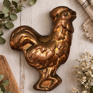 May include: A vintage copper rooster mold with intricate detailing. The mold is a warm, metallic brown color, with a textured surface. The rooster is posed in a standing position. The mold is likely used for baking or decorative purposes.