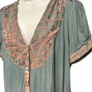 Op de afbeelding: Een vintage-stijl jurk in een zachte teal kleur, met een V-hals en korte mouwen. De jurk is versierd met ingewikkelde geborduurde panelen in een koraaltint, met bijpassende afwerking langs de halslijn, mouwen en knoopsluiting. De jurk heeft een knoopsluiting.