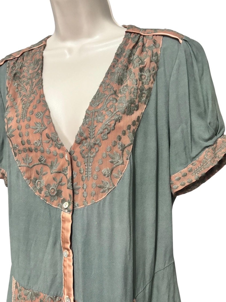 Op de afbeelding: Een vintage-stijl jurk in een zachte teal kleur, met een V-hals en korte mouwen. De jurk is versierd met ingewikkelde geborduurde panelen in een koraaltint, met bijpassende afwerking langs de halslijn, mouwen en knoopsluiting. De jurk heeft een knoopsluiting.