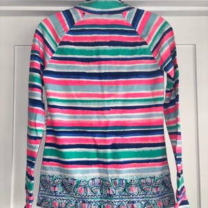 May include: A long-sleeved shirt with a vibrant striped pattern in pink, blue, green, and white. The shirt features a decorative patterned border at the hem. The shirt is hanging on a white door.