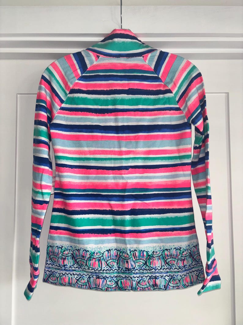 May include: A long-sleeved shirt with a vibrant striped pattern in pink, blue, green, and white. The shirt features a decorative patterned border at the hem. The shirt is hanging on a white door.