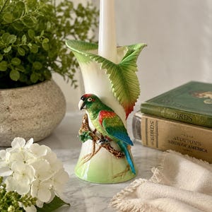 May include: A ceramic candle holder featuring a sculpted green, red, and blue parrot perched on a brown branch. The holder has a flared top rim shaped like green leaves and a white tapered candle inserted into the center of the cylindrical base.