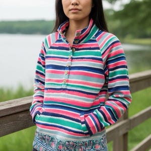 May include: A striped long-sleeve shirt with a high collar and wooden buttons. The shirt features horizontal stripes in pink, blue, green, and white. The bottom hem has a patterned design. The person is wearing black jeans.