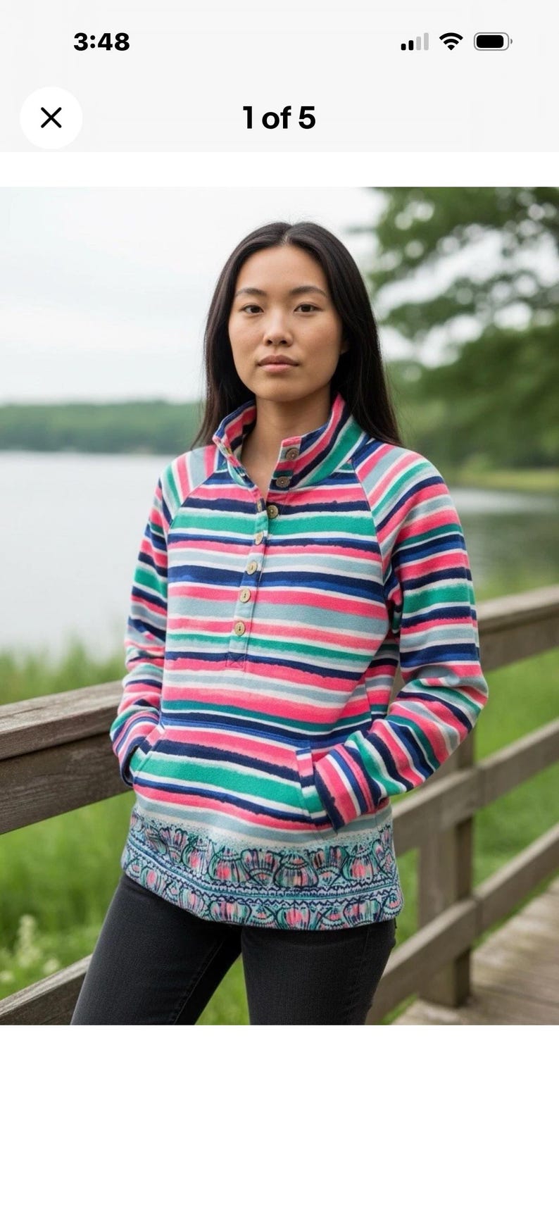 May include: A striped long-sleeve shirt with a high collar and wooden buttons. The shirt features horizontal stripes in pink, blue, green, and white. The bottom hem has a patterned design. The person is wearing black jeans.