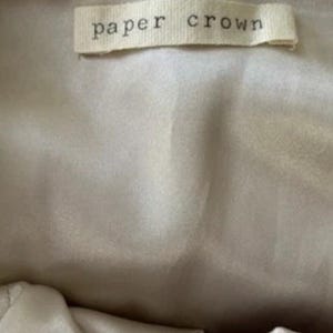 May include: A close-up of a beige fabric with a rectangular label that reads "paper crown" in a simple, sans-serif font. The label is off-white and appears to be sewn onto the fabric.