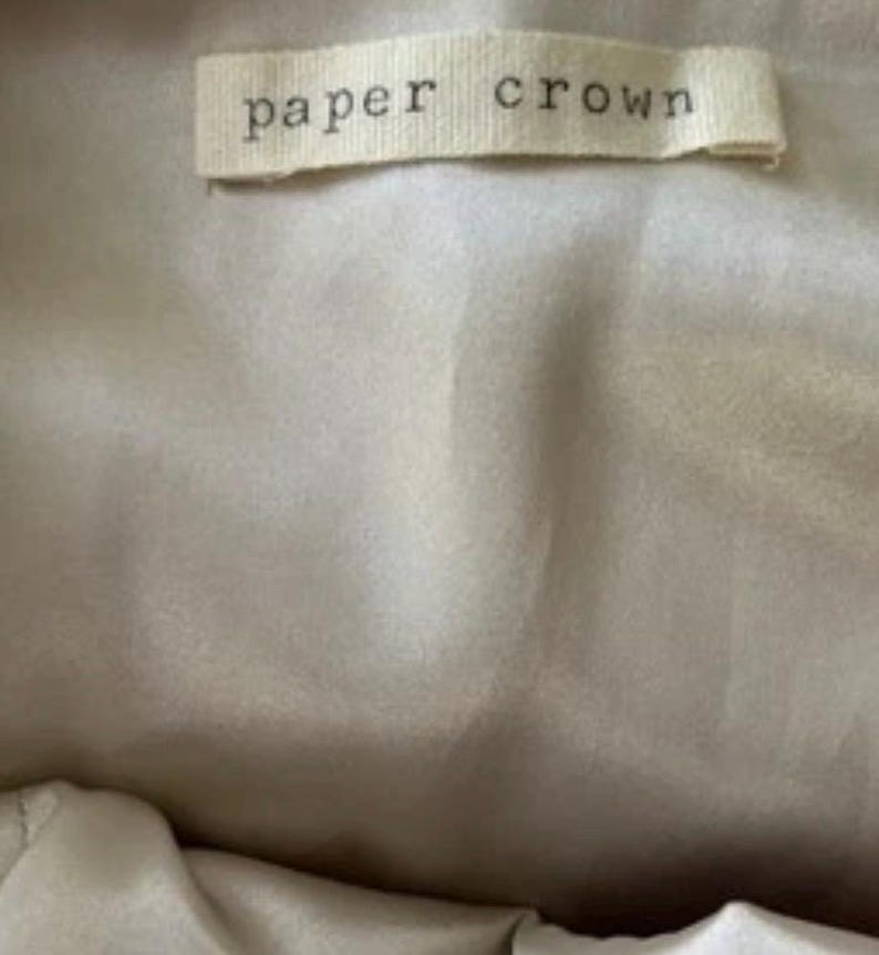 May include: A close-up of a beige fabric with a rectangular label that reads "paper crown" in a simple, sans-serif font. The label is off-white and appears to be sewn onto the fabric.
