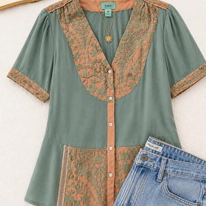 Op de afbeelding: Een saliegroene blouse met korte mouwen en een V-hals, met geborduurde details in een roestkleur. De blouse heeft een knoopsluiting aan de voorkant en wordt gecombineerd met een lichtblauwe spijkerbroek. Een gouden ketting is zichtbaar.