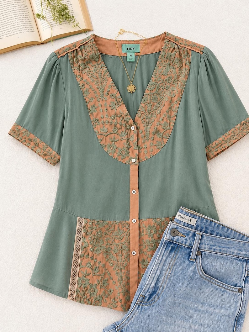 Op de afbeelding: Een saliegroene blouse met korte mouwen en een V-hals, met geborduurde details in een roestkleur. De blouse heeft een knoopsluiting aan de voorkant en wordt gecombineerd met een lichtblauwe spijkerbroek. Een gouden ketting is zichtbaar.