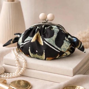 Puede incluir: Un peque&ntilde;o bolso de tela redondeado con base negra y patrones abstractos en verde y beige. El bolso cuenta con un cierre met&aacute;lico plateado de tipo boquilla rematado con dos cuentas de madera clara y laterales fruncidos.