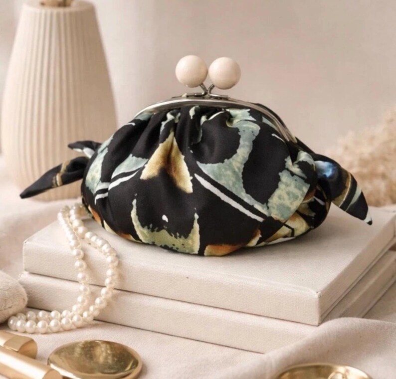 Puede incluir: Un peque&ntilde;o bolso de tela redondeado con base negra y patrones abstractos en verde y beige. El bolso cuenta con un cierre met&aacute;lico plateado de tipo boquilla rematado con dos cuentas de madera clara y laterales fruncidos.