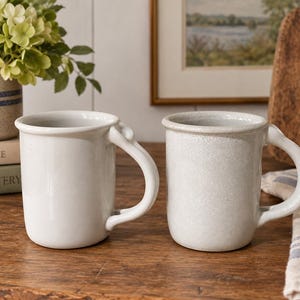 May include: Two ceramic mugs with curved handles. The left mug has a smooth, glossy white finish. The right mug features a textured, speckled light gray glaze. Both mugs have a cylindrical shape with a slightly flared rim and a rounded base.