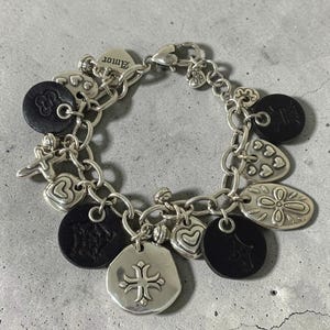 May include: Silver charm bracelet with various charms, including hearts, flowers, and a cross pendant. The bracelet also features black leather accents and the word "Amor" on a heart-shaped charm. The bracelet is on a grey surface.