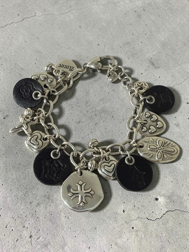May include: Silver charm bracelet with various charms, including hearts, flowers, and a cross pendant. The bracelet also features black leather accents and the word "Amor" on a heart-shaped charm. The bracelet is on a grey surface.