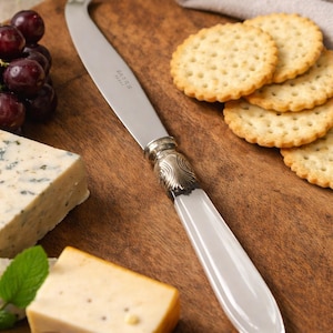 May include: A cheese knife with a silver blade and a white handle rests on a wooden cutting board. The knife is next to crackers, grapes, and various cheeses. The knife has the word "PARIS" on the blade.