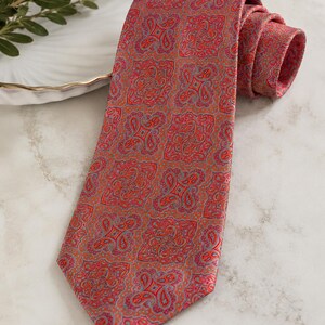 May include: A necktie featuring a repeating square pattern in shades of red, orange, and light blue. The design consists of intricate paisley-inspired motifs arranged in a grid, with the fabric rolled at the top and tapering to a pointed end.
