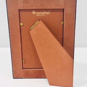 May include: Back view of a rectangular photo frame with a brown backing and stand. The frame has a black border and gold-colored metal clips. The brand name "natalini" is printed in gold, with "MADE IN ITALY" below it.