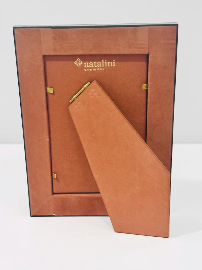 May include: Back view of a rectangular photo frame with a brown backing and stand. The frame has a black border and gold-colored metal clips. The brand name "natalini" is printed in gold, with "MADE IN ITALY" below it.