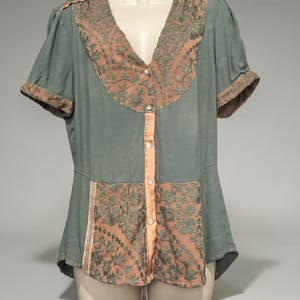 Op de afbeelding: Een vintage blouse in gedempt teal met perzikkleurige accenten. De blouse heeft een V-hals en korte mouwen met perzikafwerking. Perzikkleurige geborduurde panelen en een knoopsluiting aan de voorkant voegen details toe aan deze elegante top.