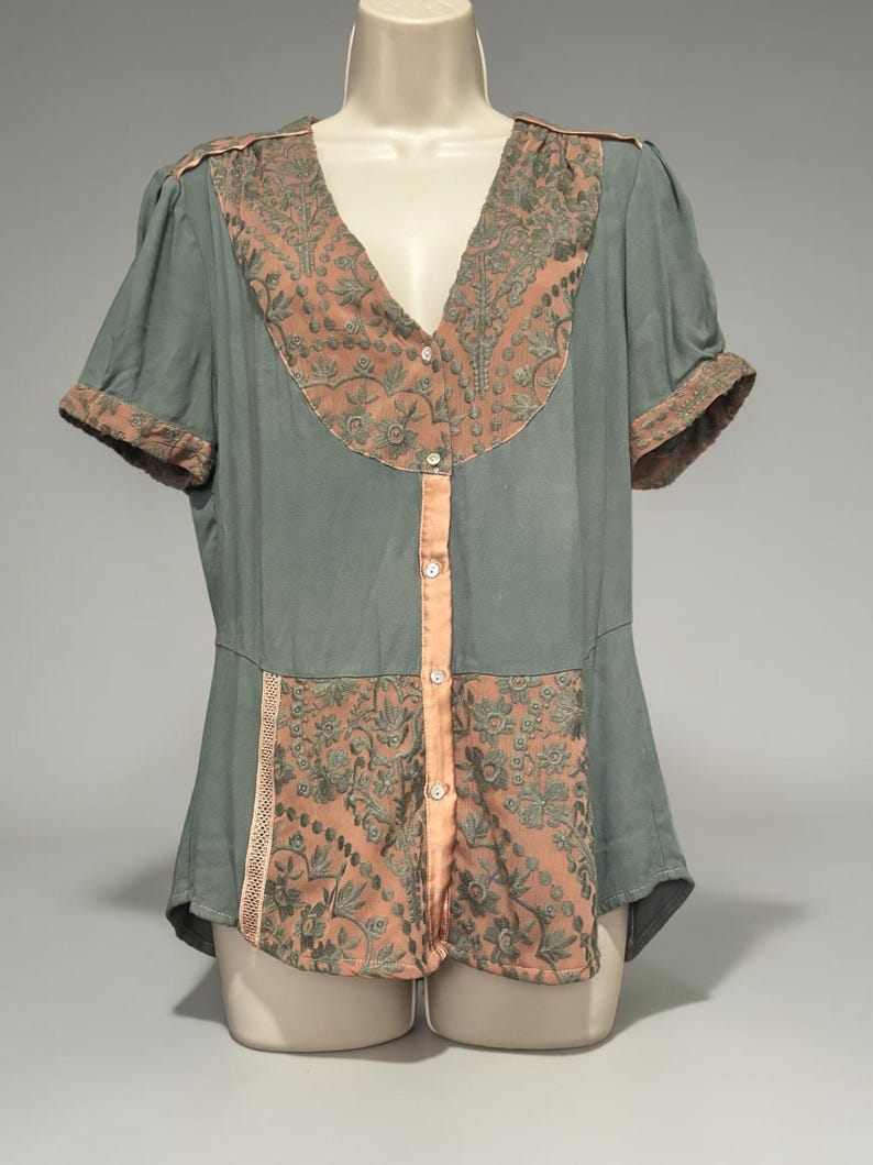 Op de afbeelding: Een vintage blouse in gedempt teal met perzikkleurige accenten. De blouse heeft een V-hals en korte mouwen met perzikafwerking. Perzikkleurige geborduurde panelen en een knoopsluiting aan de voorkant voegen details toe aan deze elegante top.