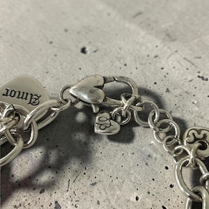 May include: Close-up of a silver bracelet featuring a heart-shaped clasp and charm. The charm is engraved with the word "Amor". The bracelet is made of interconnected oval links.