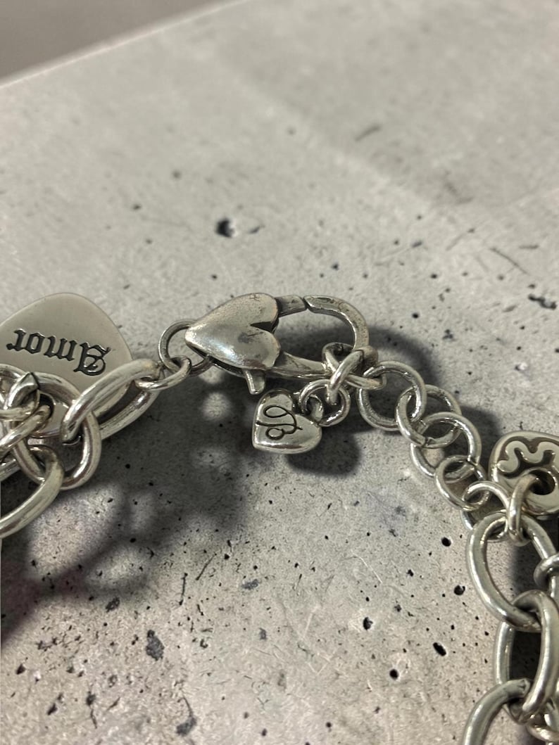 May include: Close-up of a silver bracelet featuring a heart-shaped clasp and charm. The charm is engraved with the word "Amor". The bracelet is made of interconnected oval links.