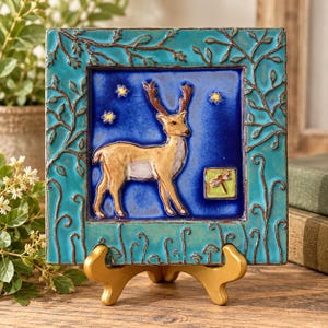 May include: A square ceramic tile featuring a central blue panel with a tan deer, yellow stars, and a small green square containing a pink dragonfly. The border is a textured turquoise frame with raised brown branch and vine details.