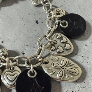 May include: Silver charm bracelet featuring a variety of charms. Charms include hearts, a four-leaf clover, and black circular charms with embossed designs. The bracelet is made of silver-toned metal and has a lobster clasp.