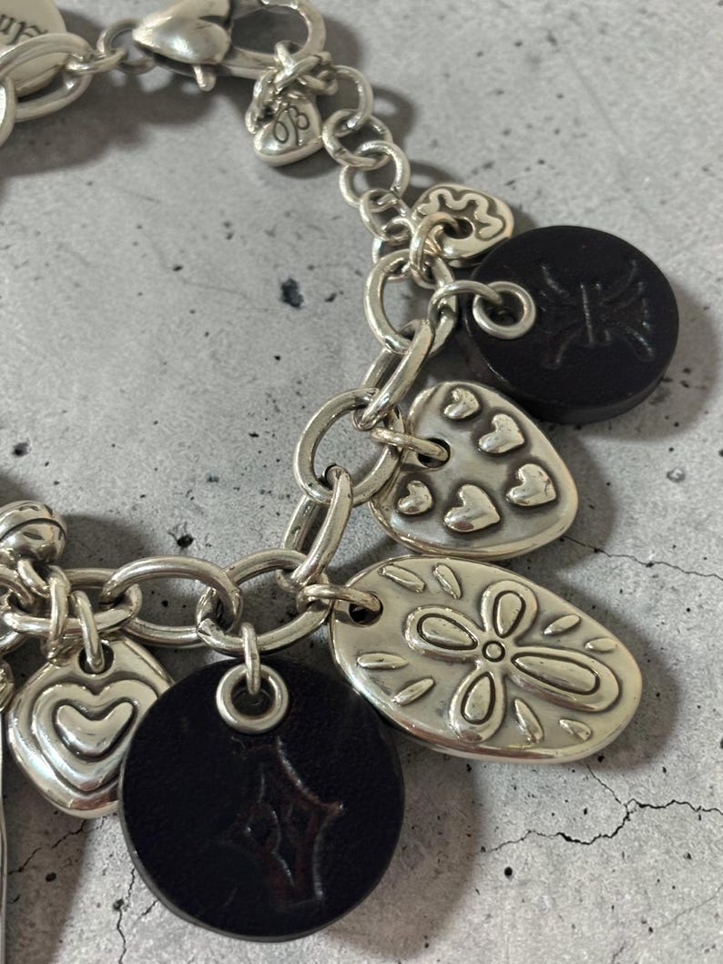 May include: Silver charm bracelet featuring a variety of charms. Charms include hearts, a four-leaf clover, and black circular charms with embossed designs. The bracelet is made of silver-toned metal and has a lobster clasp.
