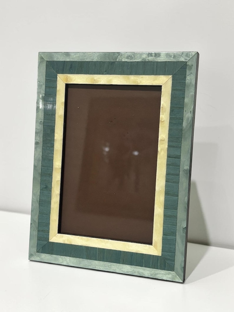 May include: Rectangular picture frame with a green and cream-colored mosaic design. The frame has a dark brown interior and is set against a white background. The frame's design features a layered effect with a light green outer border and a cream-colored inner border.