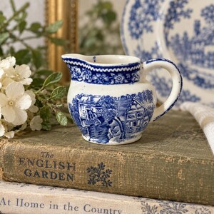 Può includere: Una piccola lattiera in ceramica con base bianca e motivi blu a trasferimento che raffigurano un paesaggio rurale con edifici e alberi. Il bordo è decorato con una fascia floreale e il manico è curvo per una presa agevole.