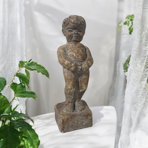 Op de afbeelding: Een bronzen beeld van een jong kind op een vierkante basis. Het kind heeft gesloten ogen en gevouwen handen. Het beeld is ongeveer 30 cm hoog en heeft een verweerde uitstraling.