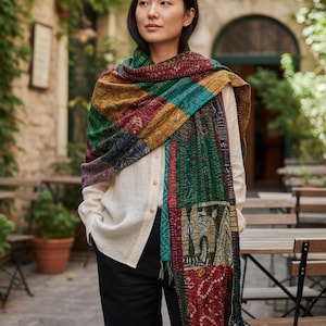 Könnte beinhalten: Eine Frau trägt einen bunten Patchwork-Schal mit verschiedenen Mustern und Farben, darunter Rot, Grün und Gold. Der Schal ist über einem weißen Hemd und einer schwarzen Hose drapiert.