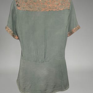 Op de afbeelding: Een saliegroene blouse met korte mouwen en een decoratief juk en manchetten. Het juk en de manchetten hebben een bloemmotief in perzikkleur. De blouse heeft een getailleerde taille en een V-hals.