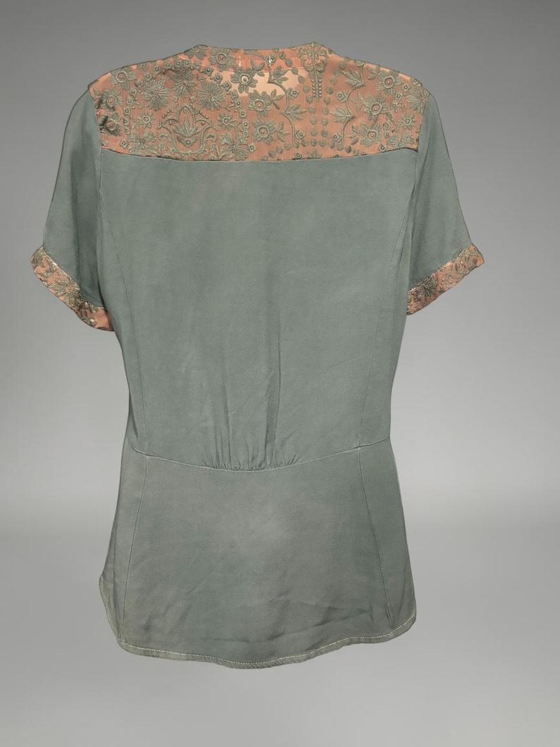 Op de afbeelding: Een saliegroene blouse met korte mouwen en een decoratief juk en manchetten. Het juk en de manchetten hebben een bloemmotief in perzikkleur. De blouse heeft een getailleerde taille en een V-hals.