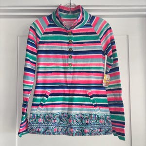 May include: A long-sleeved striped shirt with a high collar and a button-down front. The shirt features a vibrant pattern of horizontal stripes in pink, blue, green, and white. It has a decorative patterned hemline and two pockets.