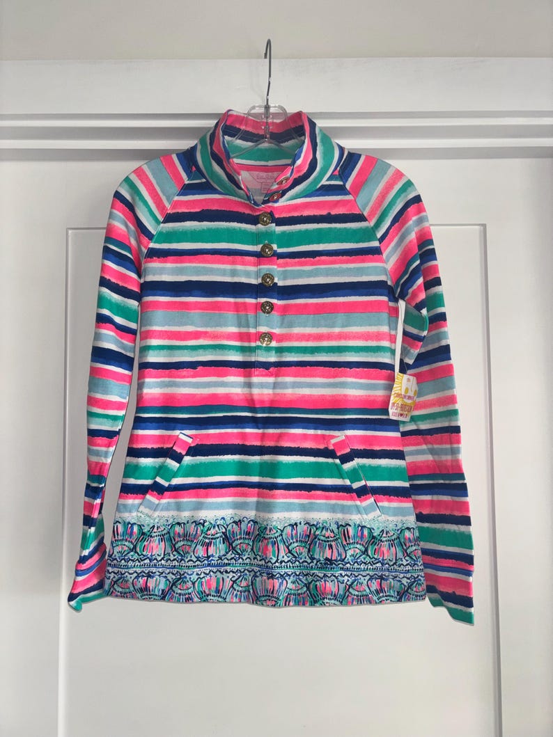 May include: A long-sleeved striped shirt with a high collar and a button-down front. The shirt features a vibrant pattern of horizontal stripes in pink, blue, green, and white. It has a decorative patterned hemline and two pockets.