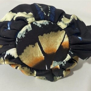 Puede incluir: Un accesorio para el cabello circular de tela con un dise&ntilde;o fruncido y plisado. El material es negro con un patr&oacute;n impreso de formas abstractas en tonos crema, bronceado y azul. La tela tiene un acabado suave y ligeramente reflectante.