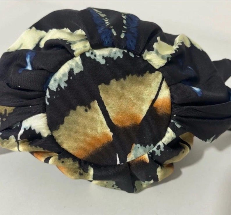Puede incluir: Un accesorio para el cabello circular de tela con un dise&ntilde;o fruncido y plisado. El material es negro con un patr&oacute;n impreso de formas abstractas en tonos crema, bronceado y azul. La tela tiene un acabado suave y ligeramente reflectante.