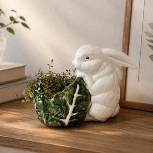 May include: A white ceramic bunny planter holding a green leaf-shaped container filled with greenery. The bunny has detailed fur texture and is positioned next to a wooden surface.