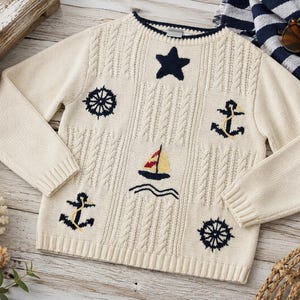 Könnte beinhalten: Cremefarbener Pullover mit maritimen Stickereien. Mit einem marineblauen Stern, Ankern, Steuerrädern und einem Segelboot. Der Pullover hat ein Zopfmuster und gerippte Bündchen und Saum.