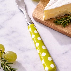 May include: A cheese fork with a stainless steel head and a bright green handle decorated with white polka dots. The fork is next to a wooden cutting board with a wedge of brie cheese, grapes, and rosemary sprigs.