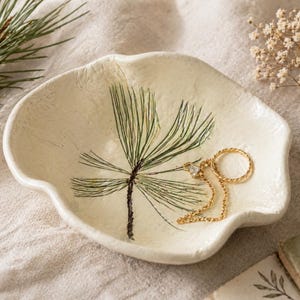 May include: A cream-colored ceramic trinket dish with a pine needle design and a gold necklace with a ring and a small charm. The dish has a wavy edge and is on a neutral-colored surface.