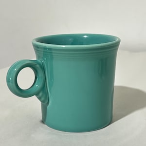 Op de afbeelding: Een effen turquoise keramische mok met een glanzende afwerking, voorzien van een cilindrisch lichaam en een cirkelvormig handvat aan de zijkant. De mok heeft een glad, uniform oppervlak met subtiele horizontale ringdetails bij de bovenrand.