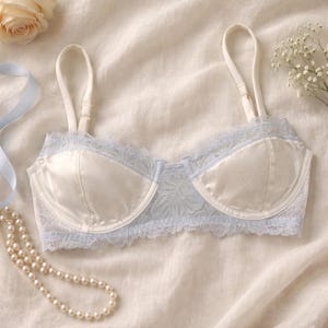 May include: A cream-coloured bra with satin cups and light blue lace trim. The bra has adjustable straps and a delicate floral pattern on the cups. The image is styled with a rose, ribbon, and pearls.