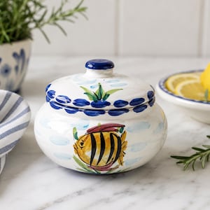 May include: A round ceramic jar with a lid, featuring a hand-painted yellow and black striped fish on a white background with light blue brushstrokes. The lid has a blue knob and a blue-painted border with green leaf accents.