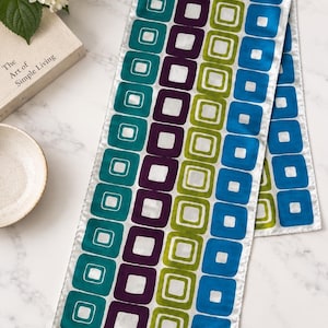May include: A rectangular scarf featuring a repeating geometric pattern of rounded squares in teal, purple, lime green, and blue. The design consists of vertical columns of these squares, each containing a smaller white square. The brand name ECHO is printed in the bottom right corner.