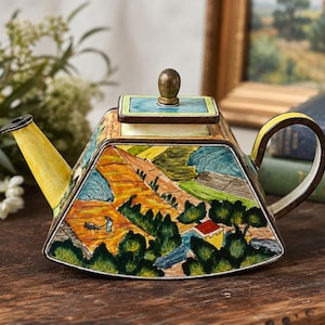 May include: A trapezoidal ceramic teapot featuring a hand-painted landscape scene with orange fields, green trees, and a small figure. The teapot has a yellow spout, a dark brown handle, and a square lid with a metallic-toned knob on top.