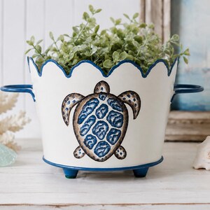 May include: A white metal planter with a scalloped top edge and two side handles. The exterior features a painted sea turtle with a blue and brown patterned shell. The planter stands on three small blue feet and contains a green leafy plant.