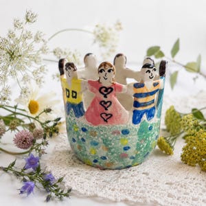 May include: A ceramic bowl featuring a ring of hand-painted figures holding hands around the rim. The figures wear colorful shirts, including yellow, pink with heart designs, and blue striped patterns. The base has a speckled green finish.