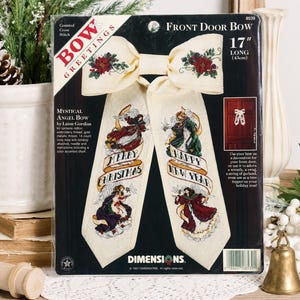May include: A Dimensions counted cross stitch kit for a 17 inch long Front Door Bow titled Mystical Angel Bow by Laine Gordon. The packaging features an ivory fabric bow with poinsettia accents, angels, and the text Merry Christmas and Happy New Year.